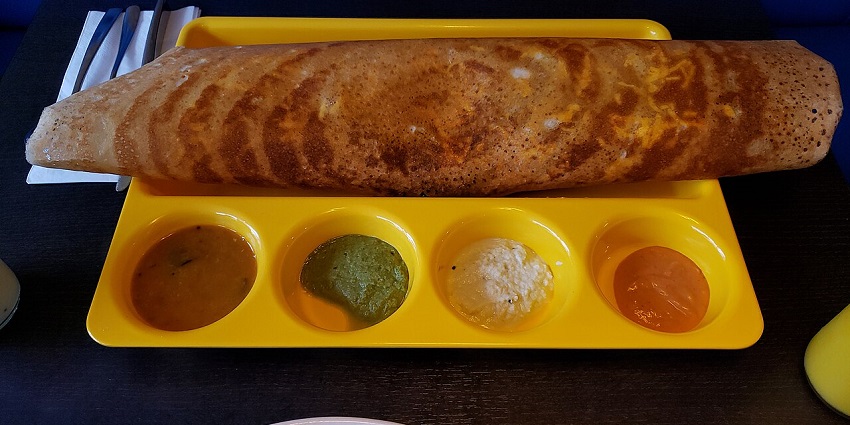 Large crispy golden dosa served with four colourful chutneys and sambar on a tray.
