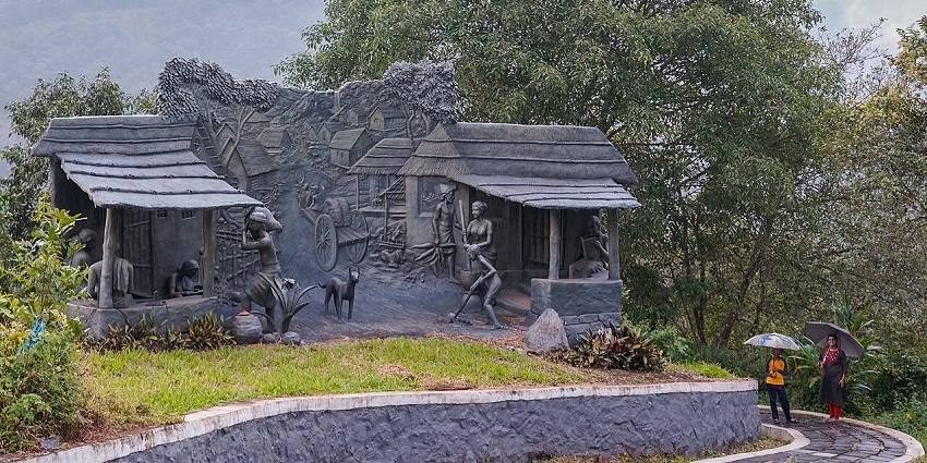 Stone sculptures depicting village life set against a green hillside and trees in Kerala.
