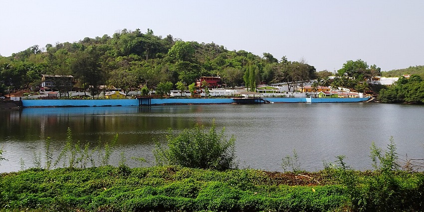 Mayem Lake Eco Park in Bicholim, with greenish, calm water body of the lake and distant buildings.
