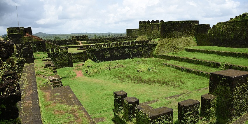 Stone walls of Mirjan Fort with lush green moss and grass under a bright blue sky, one of the forts in Karnataka.