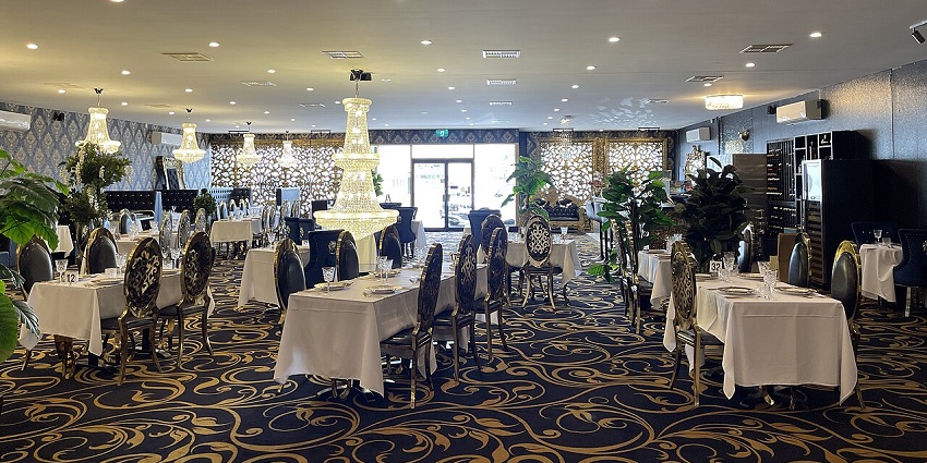 A luxurious dining room with many tables, large chandeliers, and an ornate, dark carpet.
