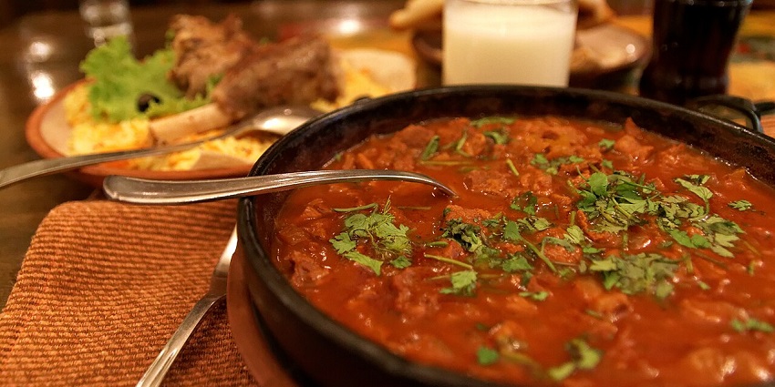 A table with a large pan of stew and two plates of meat and vegetables on it, and a bottle.
