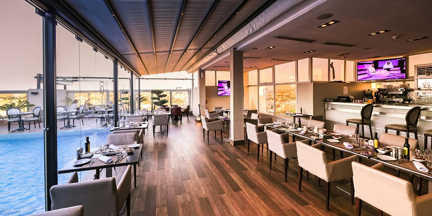 Elegant rooftop restaurant with poolside seating, glass walls, and neatly set dining tables.
