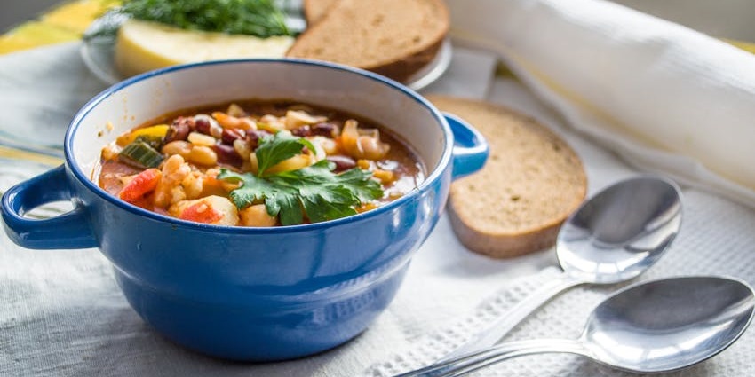 A hearty vegetable and bean soup is served in a blue bowl with a side of bread.