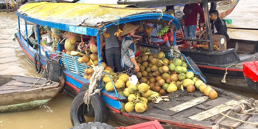 A long boat on the water is full of coconuts and pineapples, with people on board.
