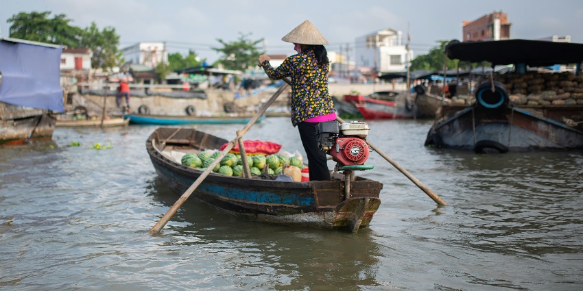 A woman in a straw hat sits in a wooden boat, paddling with watermelons for sale.
