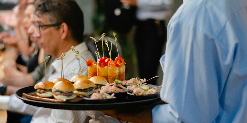 Server carrying a tray of mini burgers, prawn bites, and skewered appetizers at an event, one of the best restaurants in Orchard.