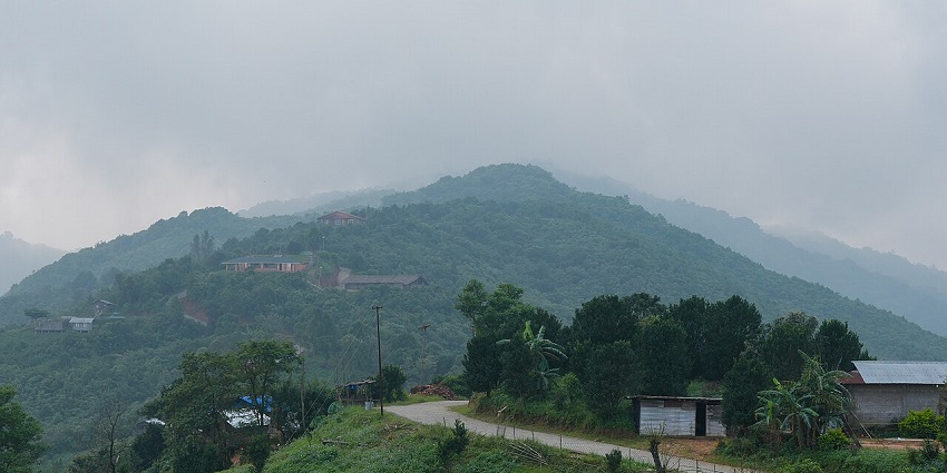 A road winds through lush green mountains with scattered houses on a foggy day.
