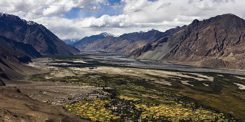 A verdant green valley with a village and fields is nestled between two large mountains.
