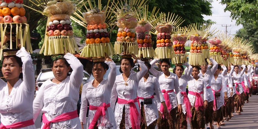 Balinese women in white attire carry tall fruit offerings during a colourful temple parade.
