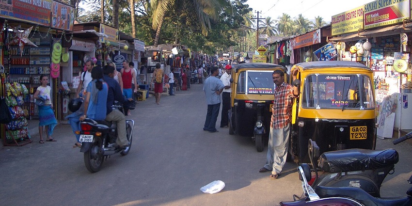 A lively street scene in India, with people, parked auto-rickshaws, and market stalls.