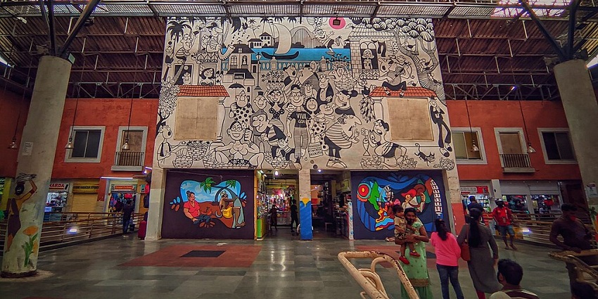 A wide shot of a busy indoor market with a large, elaborate mural on a central wall.