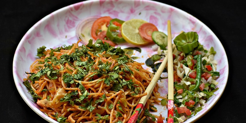 A plate of veg hakka noodles is served with slices of lemon, tomato, and onion.

