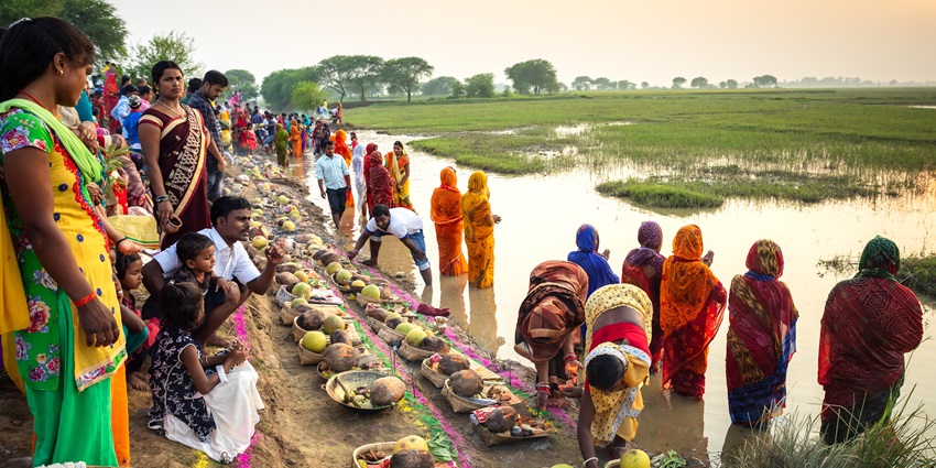 People celebrating Chhath Puja in a field with a water body reflecting the bright sun.
