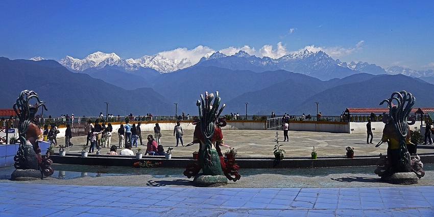 People at the glass skywalk viewpoint of Pelling with snow-covered mountains at a distance.
