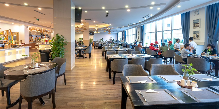 Spacious restaurant with grey chairs, wooden floors, large windows, and diners enjoying meals.
