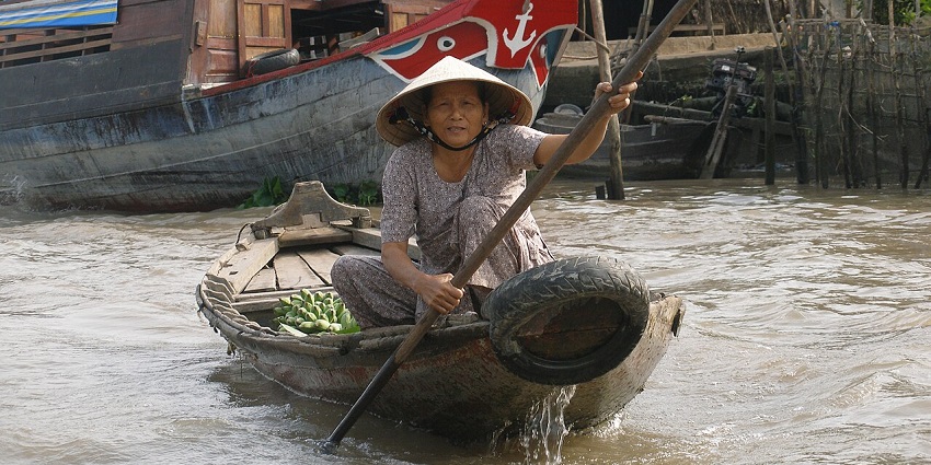 A woman in a conical hat paddles a small wooden boat on a river near a larger boat.
