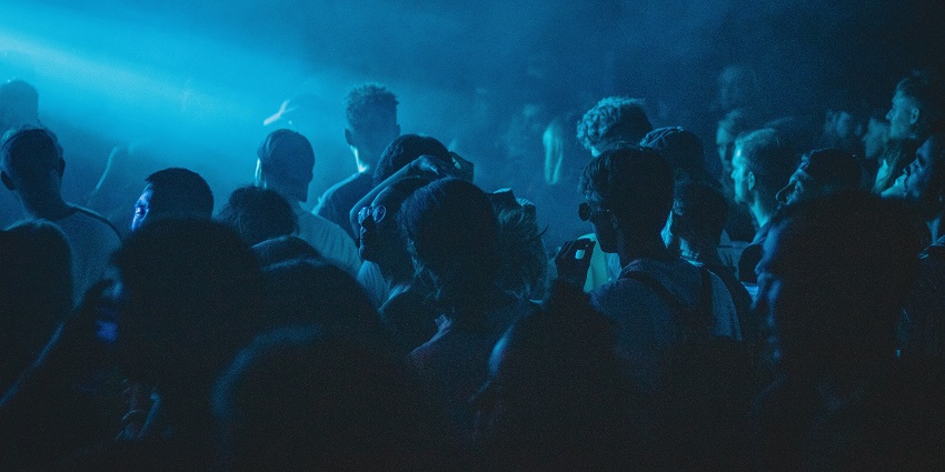 A view of a DJ takes the stage, with blue spotlights and a crowd in a nightclub.
