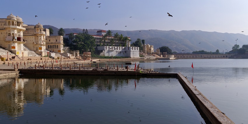 Large and calm water body of Pushkar Lake with a small red flag, buildings and birds.