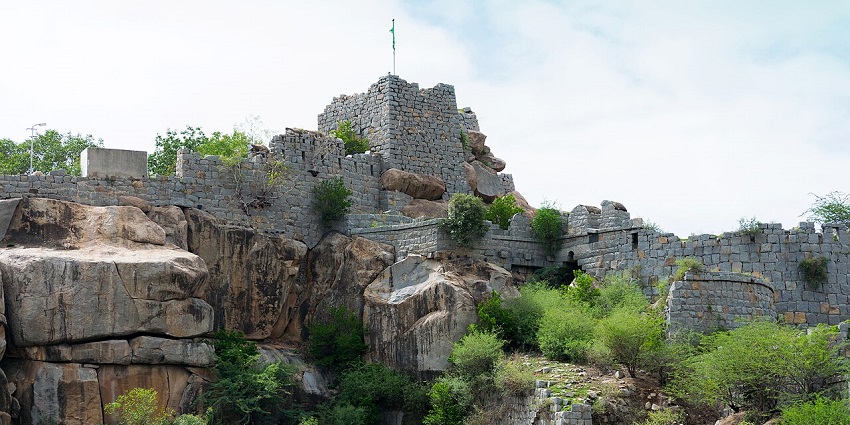 Stone walls of Raichur Fort built on a rocky hill with greenery and a flag on top under a sky.