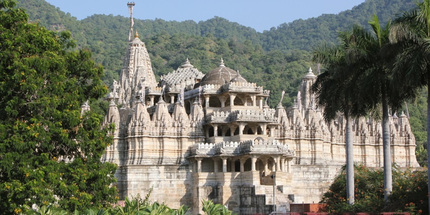 A white marble temple with multiple spires stands among trees against a backdrop of green hills, one of the hill stations in Rajasthan.