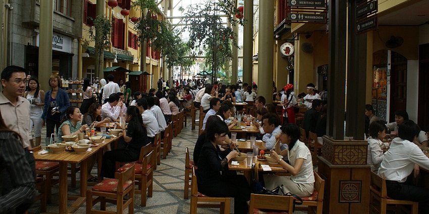 Busy dining street with wooden tables, people enjoying meals under a covered walkway, one of the best restaurants in Singapore.
