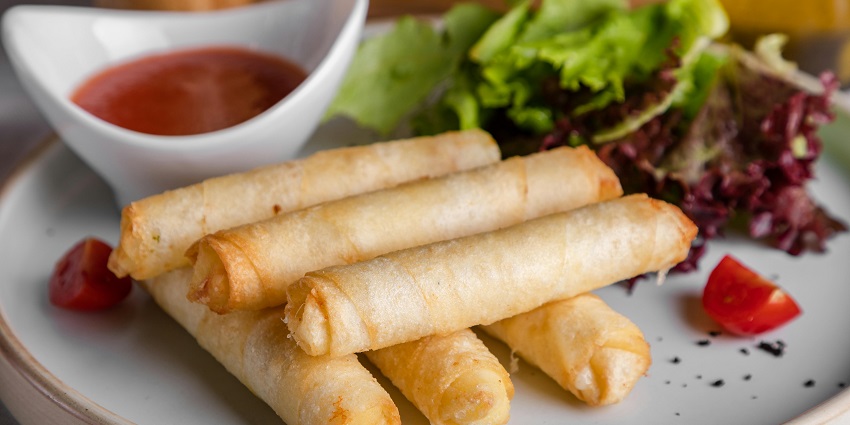 A plate of veg spring rolls is served with a small cup of chilli chutney.