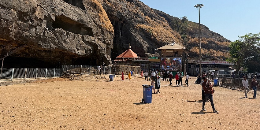 A dusty open area at the base of a large, rocky hill with a cave entrance and a small temple building.
