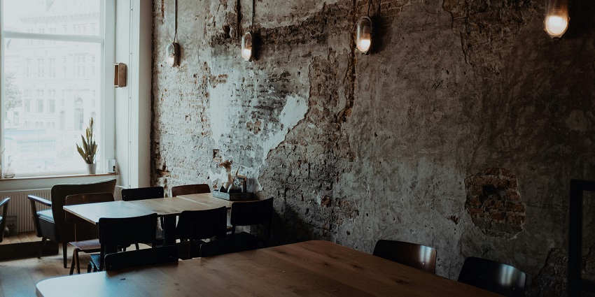 Rustic café interior with exposed brick wall, wooden tables, pendant lights, and a window.
