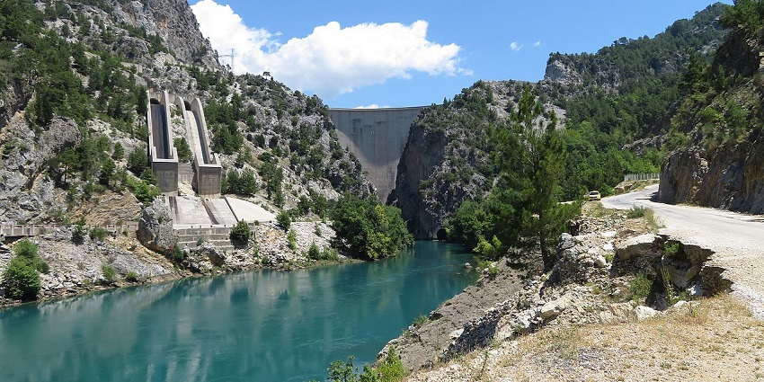 A massive concrete dam stretches between steep, rocky cliffs, with a river flowing below.
