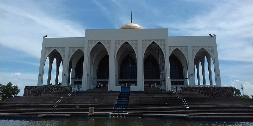 Modern mosque in Thailand with tall arched facade, golden dome, and wide front steps.
