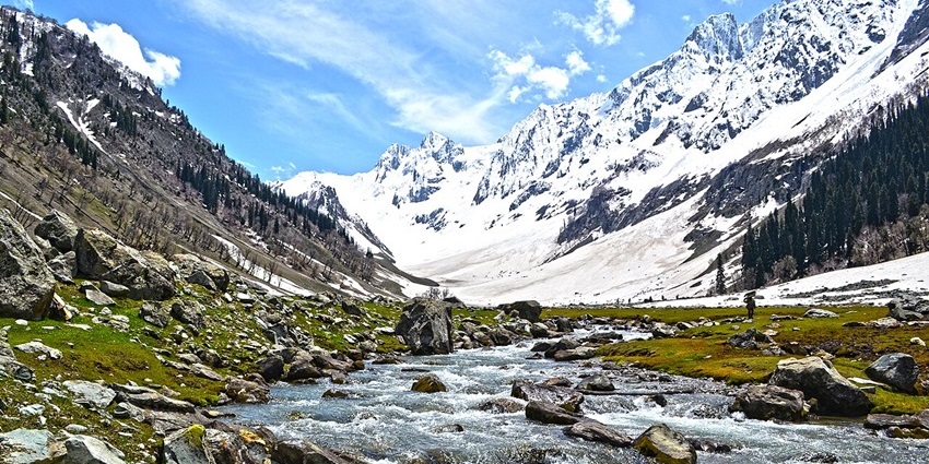 Wide stream with rocks flowing through valley of green and snowy slopes, one of the picnic places in Kashmir.