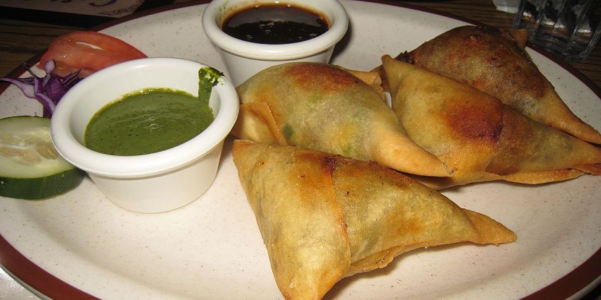 The crispy samosas are served with green and red chutney or ketchup on a metallic plate.
