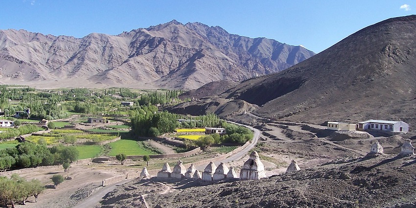 A rural valley with fields and trees, nestled between arid mountains, under a blue sky.