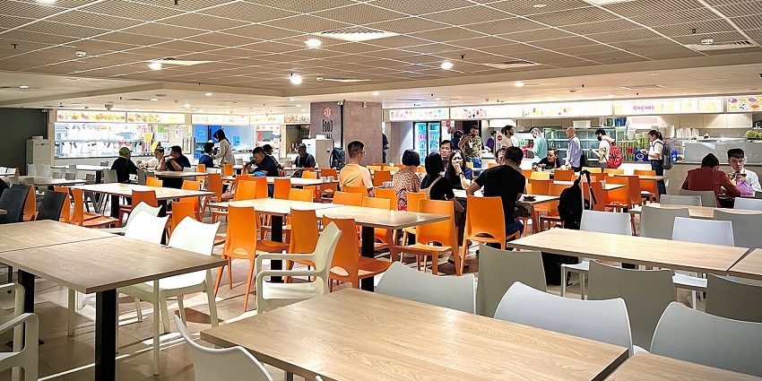 Bright food court with orange chairs, people dining and ordering from various stalls, one of the best restaurants in Singapore.
