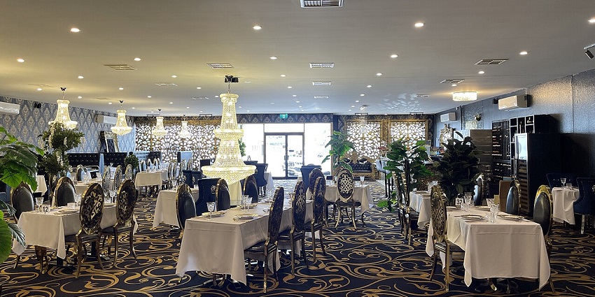 A luxurious dining room with many tables, large chandeliers, and an ornate, dark carpet.
