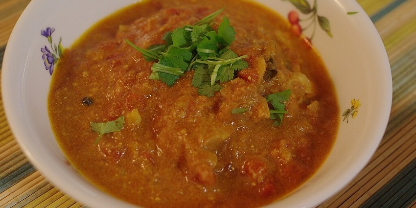 The chicken curry is served with vegetables like onions, tomatoes, and coriander in a bowl.
