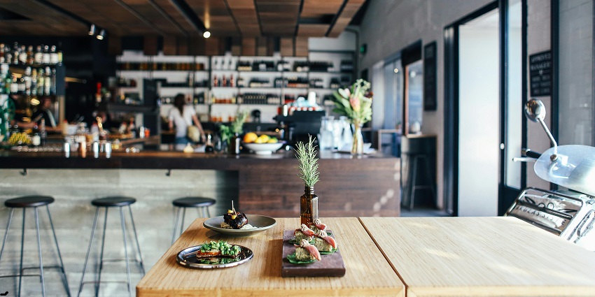 Modern café with wooden ceiling, bar counter, stools, and plated dishes on a front table.
