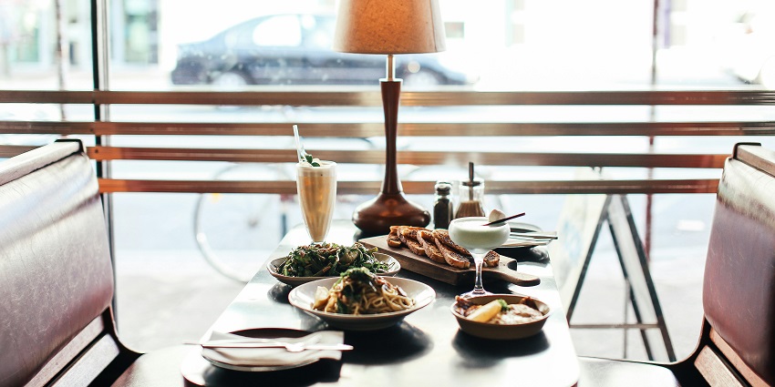 Restaurant booth table with pasta, salad, bread, drinks, and a lamp beside a large window.
