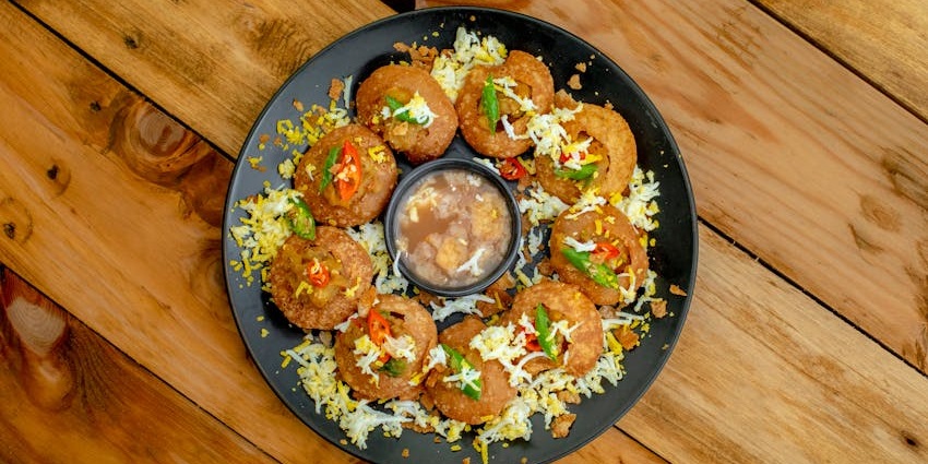 A round, black plate holds a dish of eight pani puri, garnished with spices and herbs, and a bowl of tamarind water for dipping.