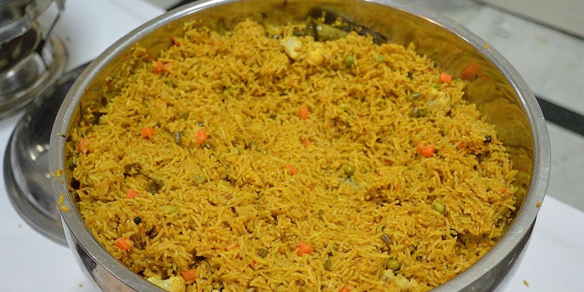 A steel pot is filled with veg biryani, featuring vegetables like Cauliflower and Peas.
