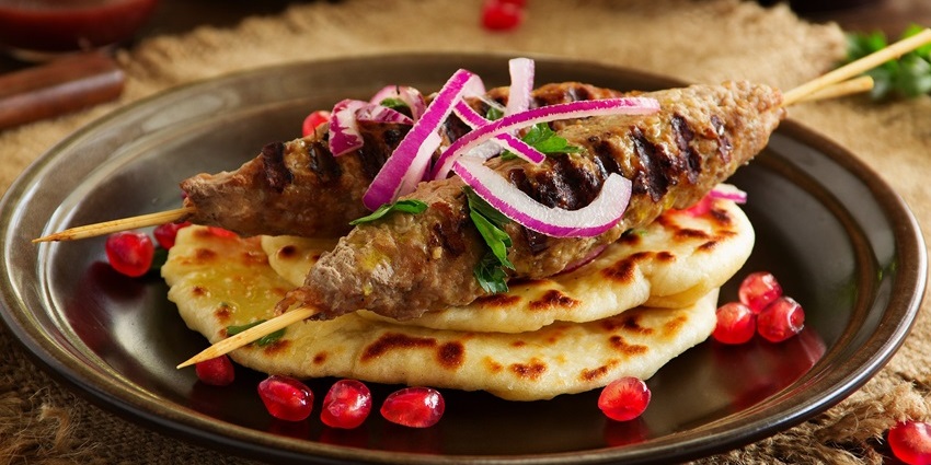 A plate of lula kabab is served with a topping of slices of onions and pomegranate seeds.