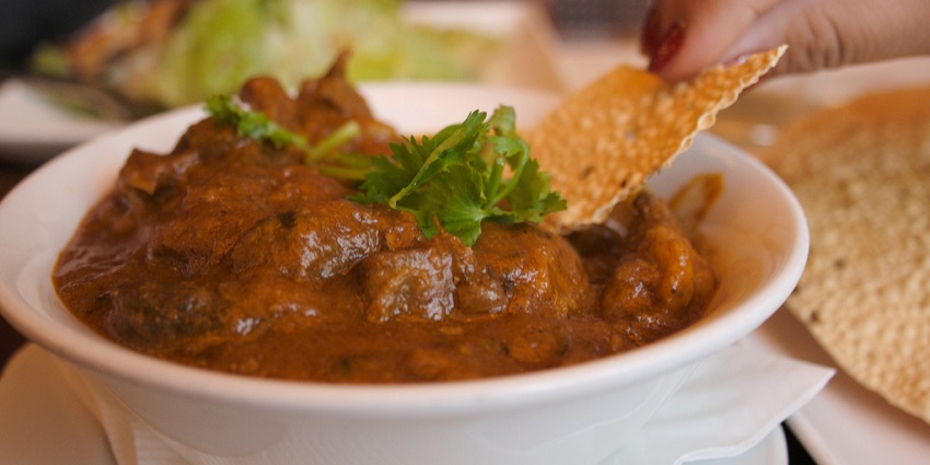 A hand holds a piece of papadum to dip into a bowl of brown curry with meat and herbs, one of the best Indian restaurants in Singapore.

