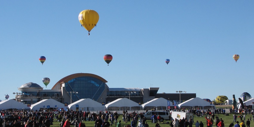 Hot air balloons at Albuquerque International Balloon Fiesta flying above people on the field with tents and a glass building.