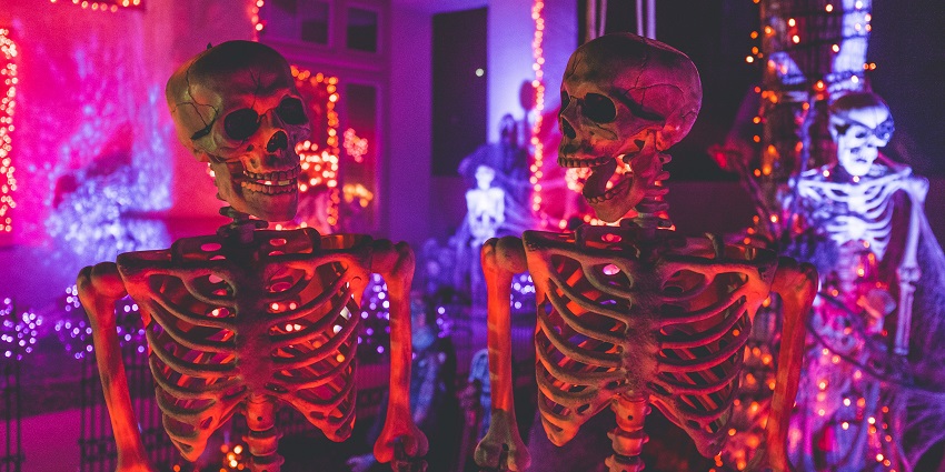 Two skeleton figures stand near a white building with string lights, creating a quirky daytime display scene.