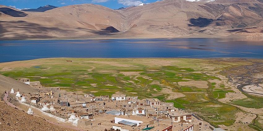 A remote village sits on a grassy plain beside a lake with mountains in the background.
