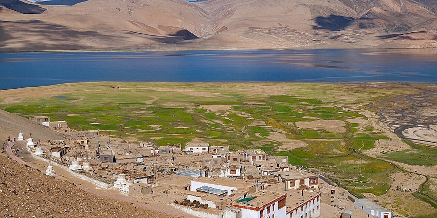 A remote village sits on a grassy plain beside a lake with mountains in the background.
