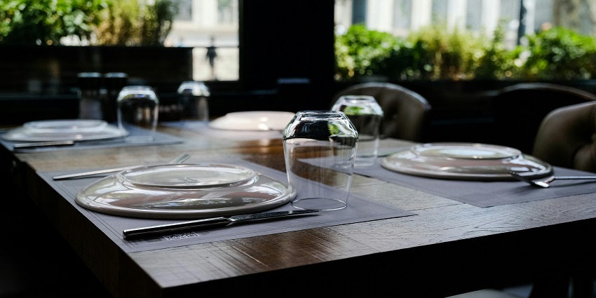 Restaurant table by window set with plates, cutlery, and glasses against a leafy backdrop.
