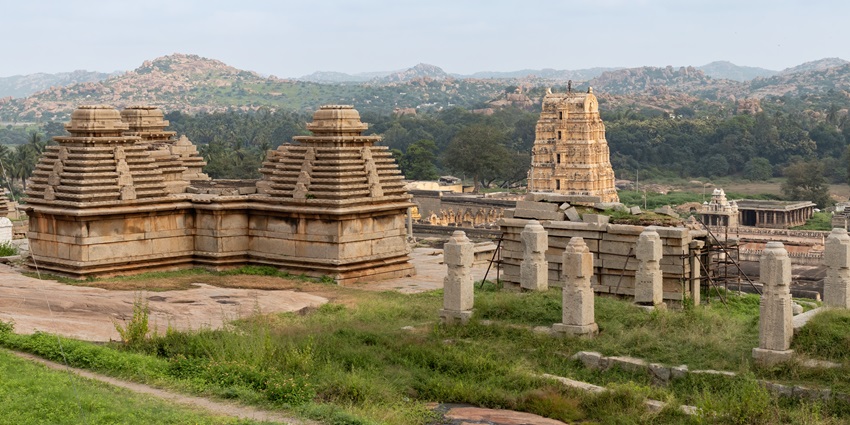 Wide-angle view of ancient temples in Karnataka amid green hills and cloudy sky.