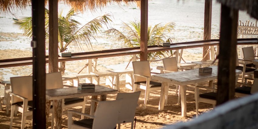 An outdoor beachside restaurant with a thatched roof, white tables, and chairs on the sand, one of the veg restaurants in Malaysia.
